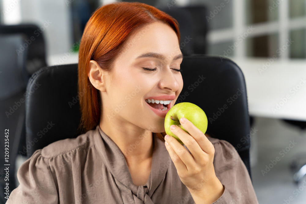 Pretty smiling ginger young woman with apple in hands sitting in the office Pretty smiling ginger young woman with apple in hands sitting in the office