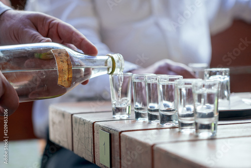 A close-up shot of a hand pouring slivovice into glass shot glasses, arranged on a wooden table in an outdoor setting, highlighting social gathering and beverage sharing. photo