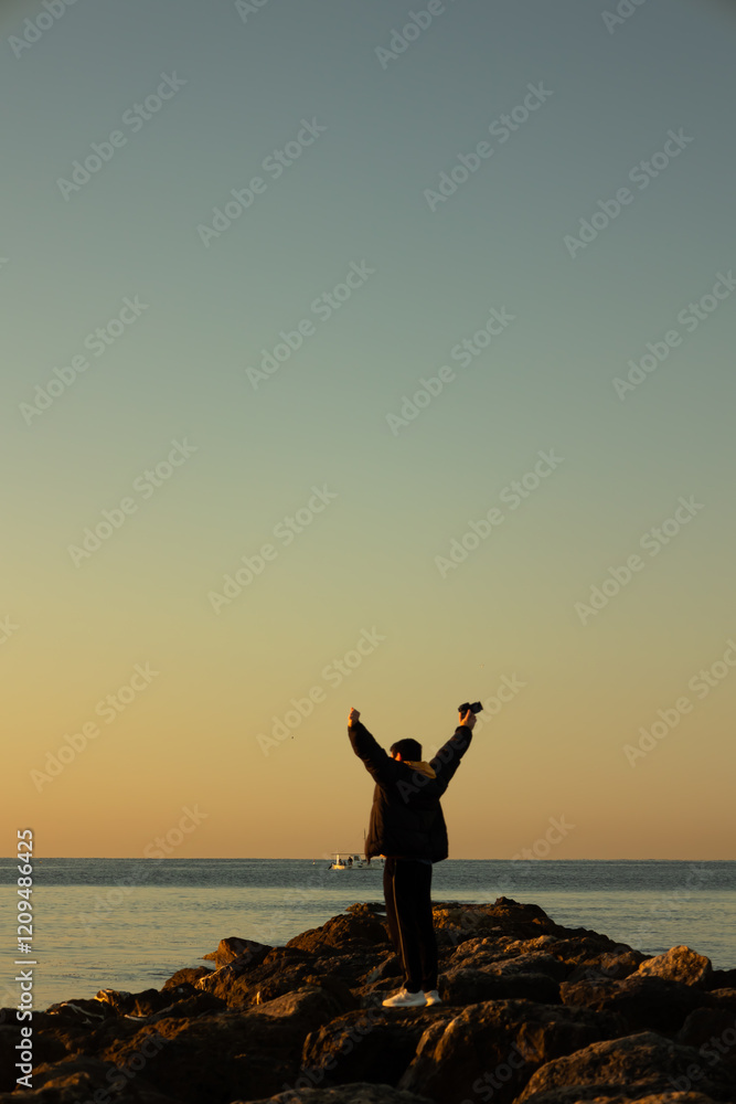 Vertical image of cheerful male photographer standing in the middle of stones raising his hands while looking at the sea on a winter morning, holding a camera in his hand. Lifestyle Vertical image of cheerful male photographer standing in the middle of stones raising his hands while looking at the sea on a winter morning, holding a camera in his hand. Lifestyle