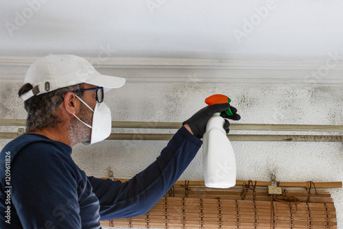 A man wearing a cap and a mask cleans mould from walls and ceilings with a sprayer. Humidity and condensation problem in homes.