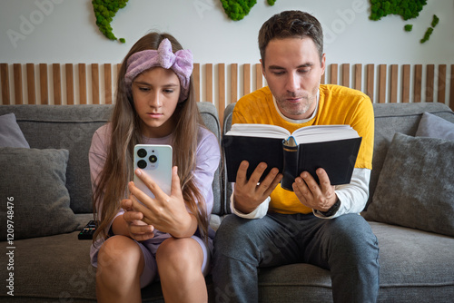 Father and daughter are sitting on the couch in the living room. girl is looking at the mobile phone, the man is reading a book. The generation x y z difference. offline and online communication photo