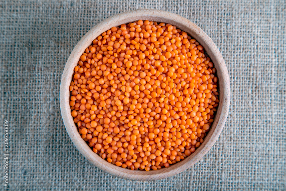 A wooden bowl with red lentils grains on a burlap. Top view of dried orange lentils inside the bowl. A wooden bowl with red lentils grains on a burlap. Top view of dried orange lentils inside the bowl.