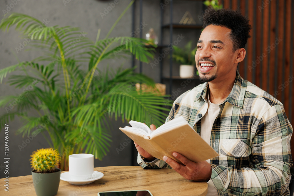Young smiling minded smart man of African American ethnicity wear casual clothes reading novel book or diary sit alone at table in coffee shop cafe relax rest in restaurant during free time indoors. Young smiling minded smart man of African American ethnicity wear casual clothes reading novel book or diary sit alone at table in coffee shop cafe relax rest in restaurant during free time indoors.