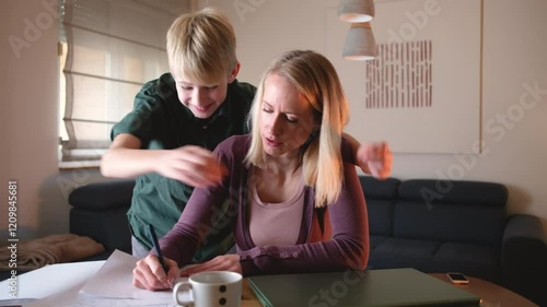 
A mother is working from home on her laptop in the living room when her son comes up and embraces her. She pauses her work to share a loving hug with him.