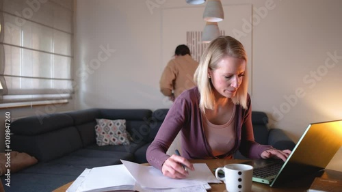 A mother works from home on her laptop in the living room, trying to focus on her tasks, while yelling at her children to calm down as they play energetically in the background.