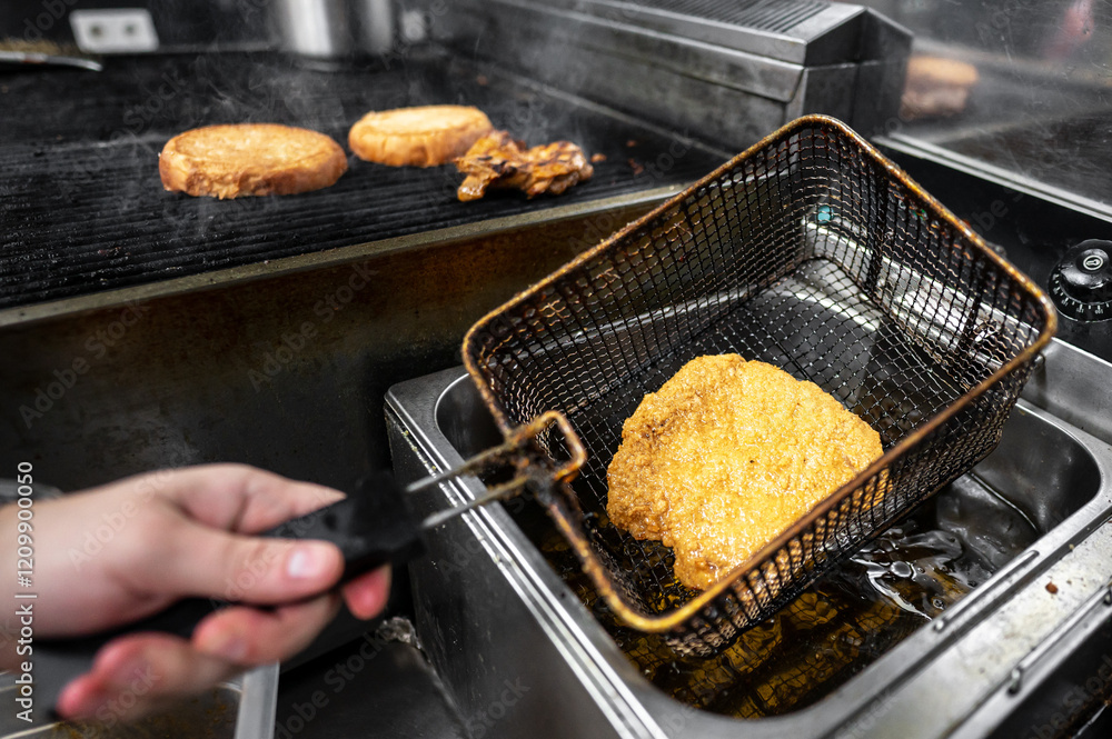 A hand lifting a golden-brown fried food item from a deep fryer, with toasted bread cooking on a grill in the background. The scene captures the bustling atmosphere of a commercial kitchen. A hand lifting a golden-brown fried food item from a deep fryer, with toasted bread cooking on a grill in the background. The scene captures the bustling atmosphere of a commercial kitchen.