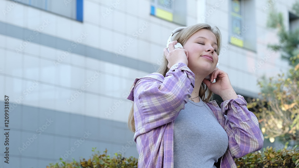 Fototapeta premium A young woman listens to music on her headphones in an outdoor urban setting, smiling softly. This photo showcases casual confidence and the joy of everyday moments.