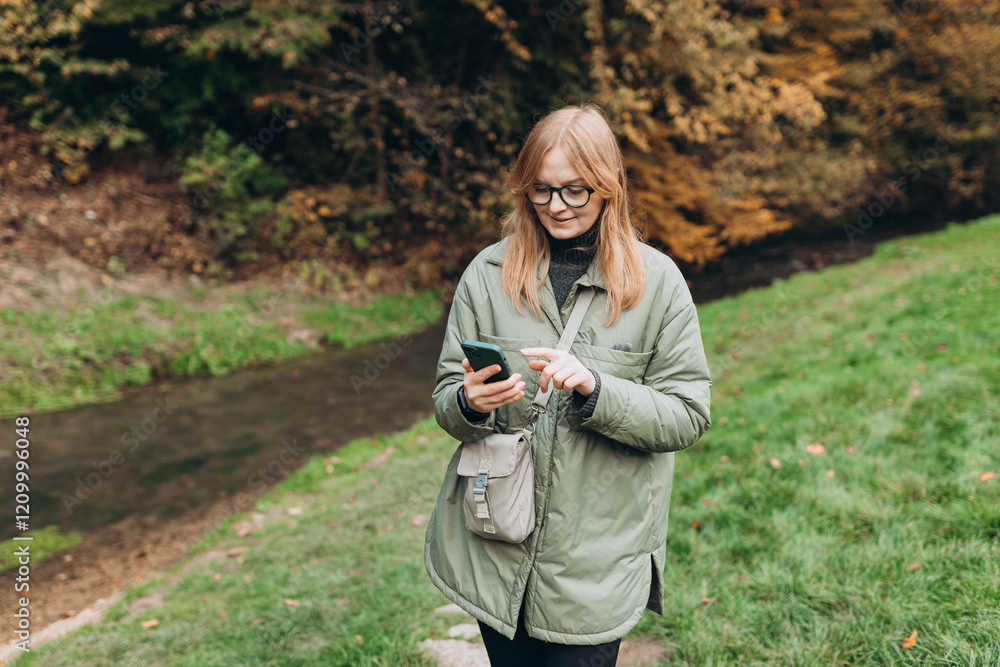 30s Girl tourist walking in forest and using smartphone. Young female hiking on the road in nature. Beautiful blonde woman walking on autumn landscape in sport clothes. High quality photo