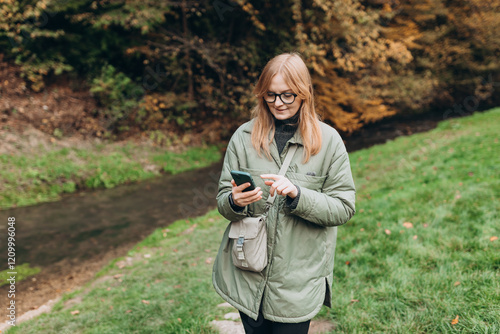 Obraz na płótnie 30s Girl tourist walking in forest and using smartphone. Young female hiking on the road in nature. Beautiful blonde woman walking on autumn landscape in sport clothes. High quality photo w salonie