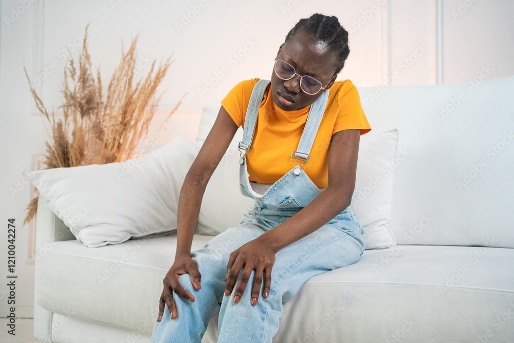 Obraz premium Young black woman wearing casual clothes sits on a white sofa, experiencing discomfort and holding her knees due to pain, possibly from an injury or medical condition
