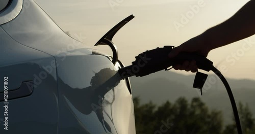Unrecognizable businessman plugging electric car from charging station. Senior plugging in power cord to electric car at cinematic sunset. Businessman charging electric car at outdoor charging station