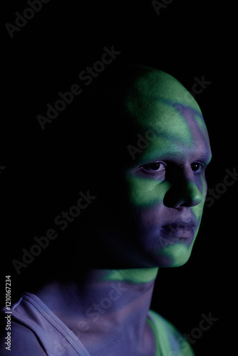 Vertical portrait of emotionless bald man looking away with sorrowful stare while standing in blue light of projector in darkness, copy space photo