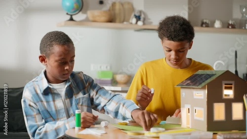 Two brothers work on school science project in cozy room. Younger assembles with glue, older cuts paper. Solar-powered cardboard house model glows nearby. Concept of teamwork and renewable energy