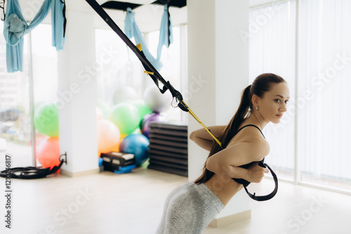 Side view portrait of muscular beautiful woman wearing sportswear squatting with fitness straps in the gym, using trx for her body workout. Indoor shot against white wall background photo