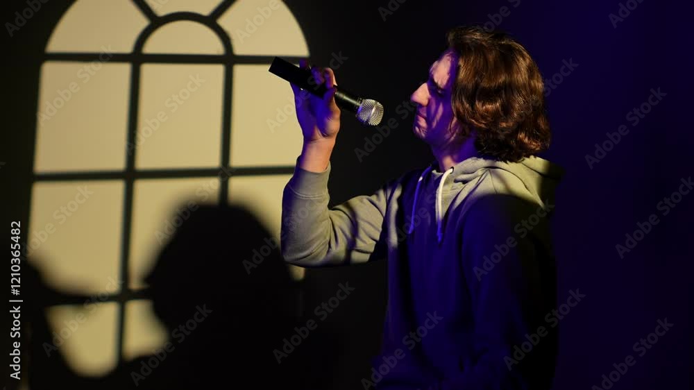 Portrait of man in blue neon singing in karaoke microphone, standing in spotlight isolated on dark wall with window pattern studio background. Portrait of man in blue neon singing in karaoke microphone, standing in spotlight isolated on dark wall with window pattern studio background.