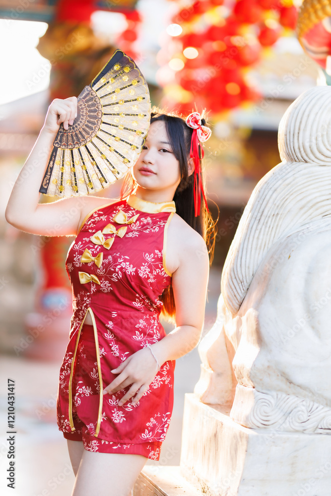 Portrait beautiful cute smiles Asian young woman wearing red traditional Chinese cheongsam decoration and holding a Chinese Fanning for Chinese New Year Festival at Chinese shrine in Thailand