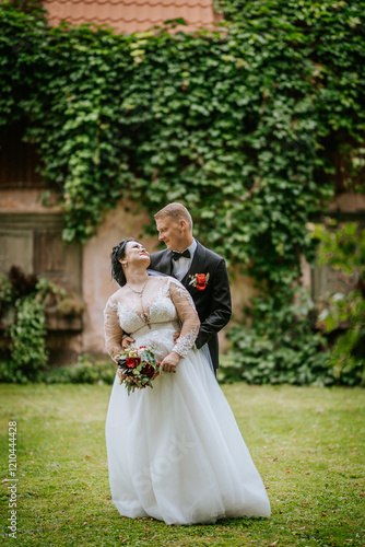 A bride in a white gown and groom in a suit share a romantic moment outdoors, standing against a green ivy-covered wall, holding a bouquet. photo