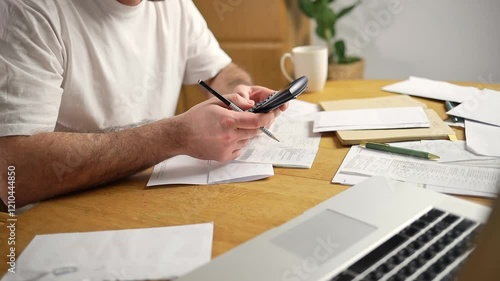 A man is sitting in a kitchen checking bills on a computer. The table is cluttered with a calculator, papers, invoices, and letters, emphasizing financial tasks and household management.