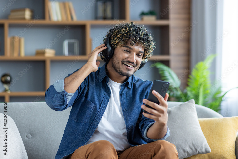 Young man sitting at home on sofa in living room, using app to listen to music online on phone. Student in headphones smiling watching online videos, and social media. Young man sitting at home on sofa in living room, using app to listen to music online on phone. Student in headphones smiling watching online videos, and social media.