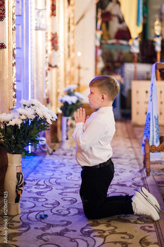 A young boy, dressed in formal attire, kneels in prayer before a religious icon inside a church. White flowers are visible in a vase nearby. photo