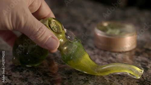 Person sets a marijuana pipe down on a granite countertop, with weed grinder in background.  Close up view of drug paraphernalia.