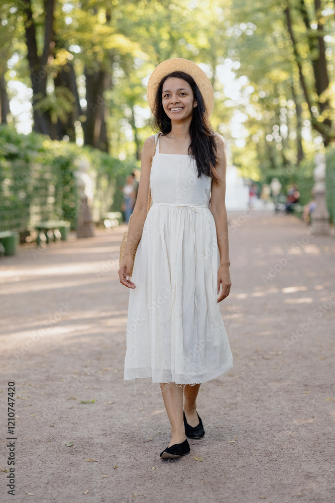 carefree African American woman in white dress and straw hat walks in park and poses against background of green hedge. daytime for relaxation. beautiful female fashion model, sunlight summer time carefree African American woman in white dress and straw hat walks in park and poses against background of green hedge. daytime for relaxation. beautiful female fashion model, sunlight summer time