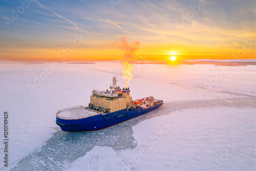 Large blue icebreaker is floating in ice, with sky behind it painted in shades of orange as sun sets in background.
