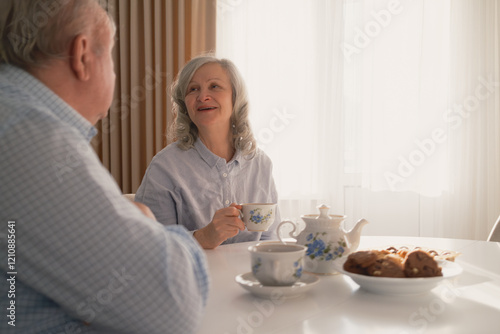 An elderly couple sits at a bright table, enjoying tea and conversation, showcasing warmth and companionship in a peaceful home environment, surrounded by beautifully crafted tableware and sunlight. photo