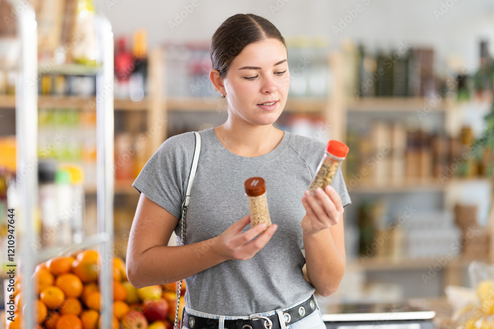 Obraz premium In healthy eating store, young woman buyer choosing spices items. Clients look package of spices bottle, inspect product and examines contents of package, compare paprika and chopped red pepper.