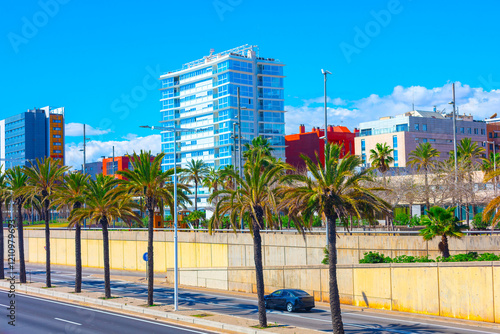 Obraz na płótnie Modern Barcelona street scene featuring palm trees, a clear blue sky, and colorful contemporary buildings, including a high-rise with reflective glass windows, alongside a road with minimal traffic w salonie