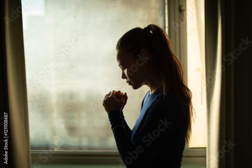Φωτογραφία Young woman with hands folded praying to god next to window