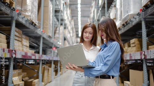In a bustling warehouse filled with shelves of products, two women share insights and strategies while working on a laptop computer, demonstrating teamwork and efficiency in their tasks.
