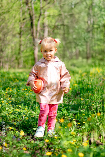 A little girl of 2-3 years old stays in the forest and enjoys nature. Background of a green forest. The girl holds tightly in her hand an orange jar in which she collects the flowers she likes.