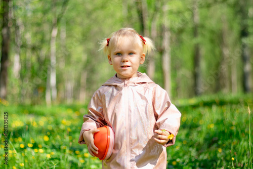 Obraz na płótnie A little girl of 2-3 years old stays in the forest and enjoys nature. Background of a green forest. The girl holds tightly in her hand an orange jar in which she collects the flowers she likes. w salonie
