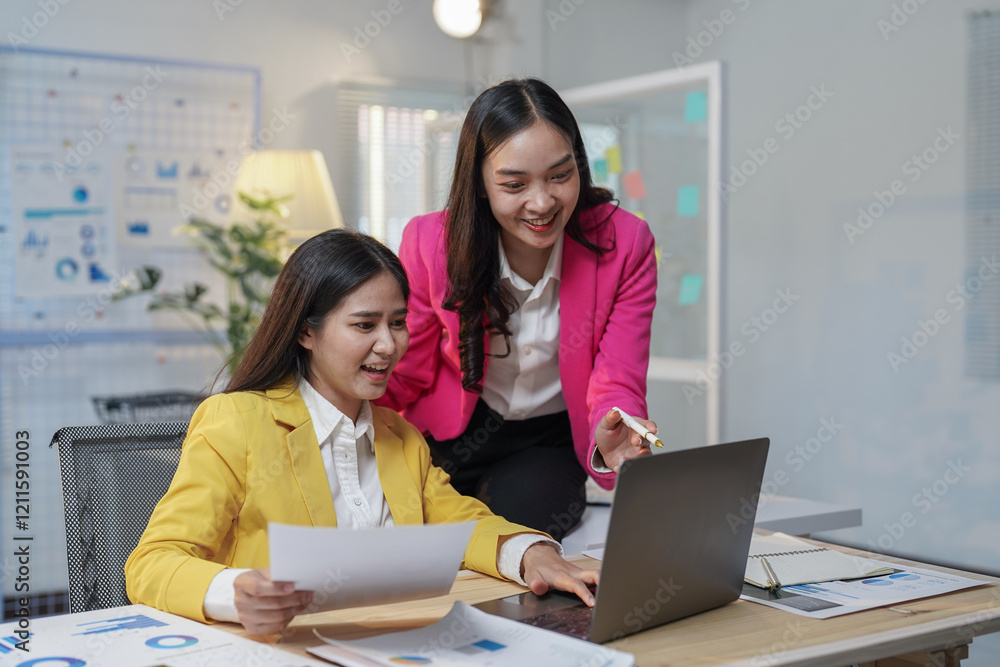 Obraz premium Two happy businesswomen collaborating in the office, using a laptop while discussing their project. Analyzing financial charts and exchanging smiles, they embody teamwork and professionalism