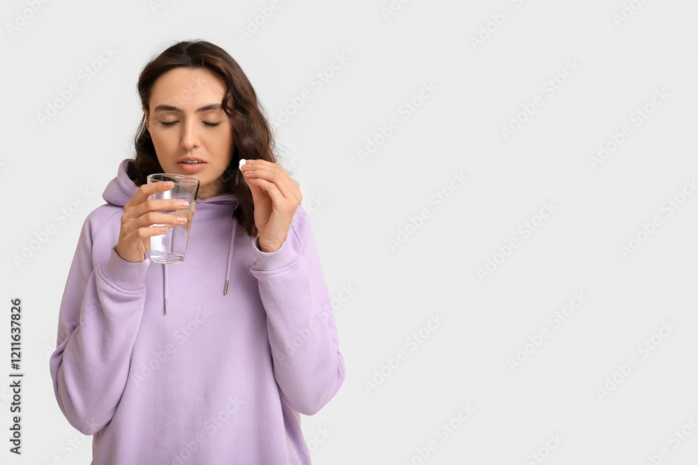 Sick young woman with glass of water taking pills on light background