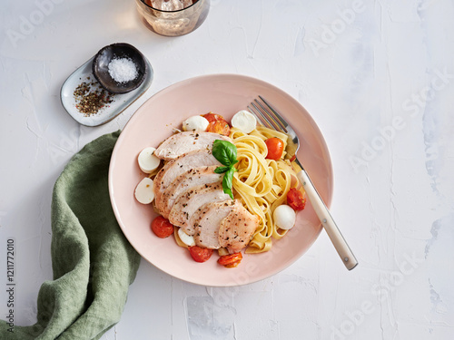 Enjoy a flavorful pasta dish featuring grilled chicken slices, fresh tomatoes, and mozzarella balls, presented in a soft pink bowl. Perfect for a relaxed meal