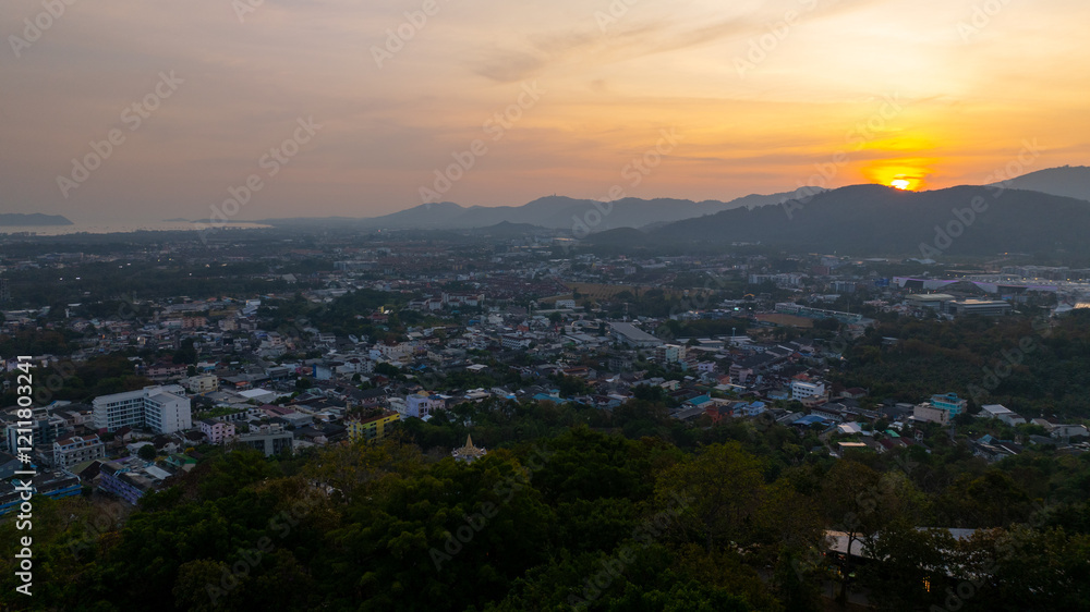 Obraz premium A scenic view of a town nestled among lush green mountains at sunset, with a temple structure perched on the hillside and a vibrant orange sky creating a warm, tranquil atmosphere.