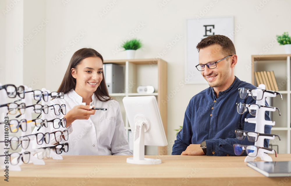 Fototapeta premium Young smiling female optician doctor helping man patient to choose new glasses after eye examination sitting on workplace in medical clinic for vision correction. Ophthalmology and eye health concept