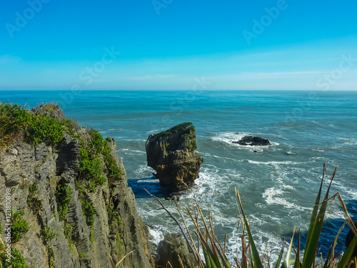 Dramatic sea stack rises from Pacific ocean along Pancake Rocks and Blowholes walking trail. Layered rock formations and native plants in Paparoa National Park. Wild beauty of New Zealand South Island photo