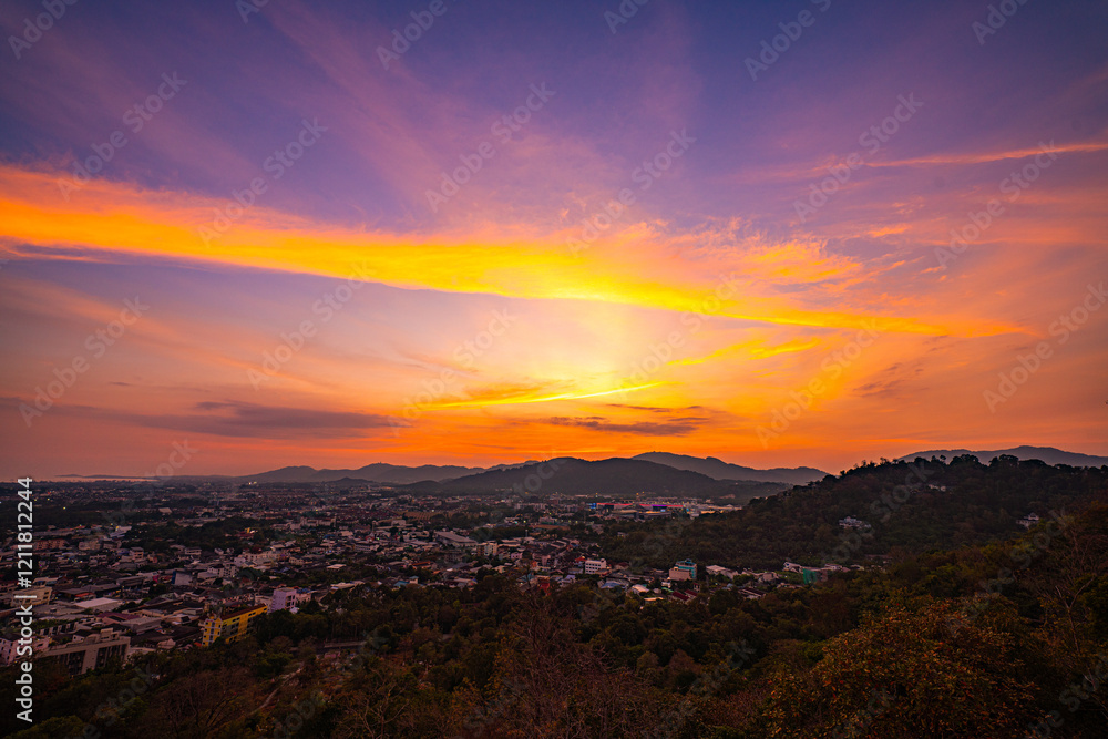 Obraz premium A scenic view of a town nestled among lush green mountains at sunset, with a temple structure perched on the hillside and a vibrant orange sky creating a warm, tranquil atmosphere.