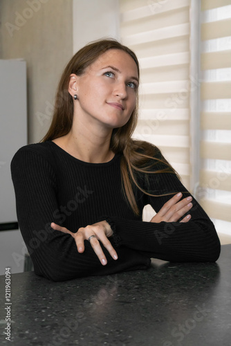 Young woman wearing a black dress is sitting at the kitchen counter, leaning her head on her hand and looking away dreamily, lost in thought. Vertical photo