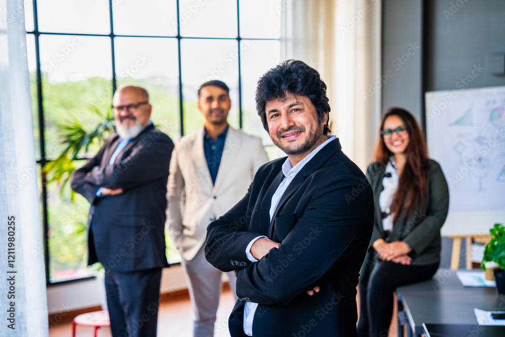 Confident Indian business leader standing with folded hands, colleagues out of focus in the office Confident Indian business leader standing with folded hands, colleagues out of focus in the office