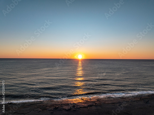 Aerial view of the setting sun touching the horizon line with its reflection in the sea, ocean, with small waves