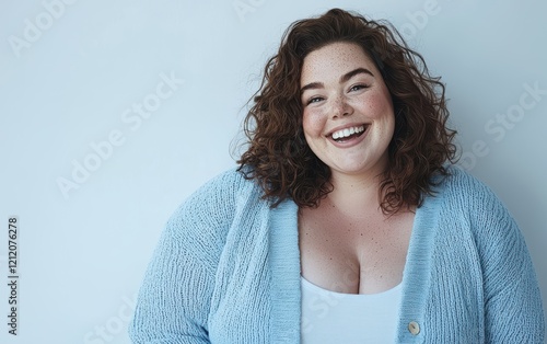 A happy full-figured woman with soft freckles and a joyful smile, dressed in a pastel blue cardigan, posed on a minimalistic white surface