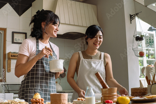 Two Asian women enjoying a baking session in a cozy kitchen. One is cutting waffles while the other pours milk into a bowl. The wooden table displays finished dishes like waffles and pizza photo