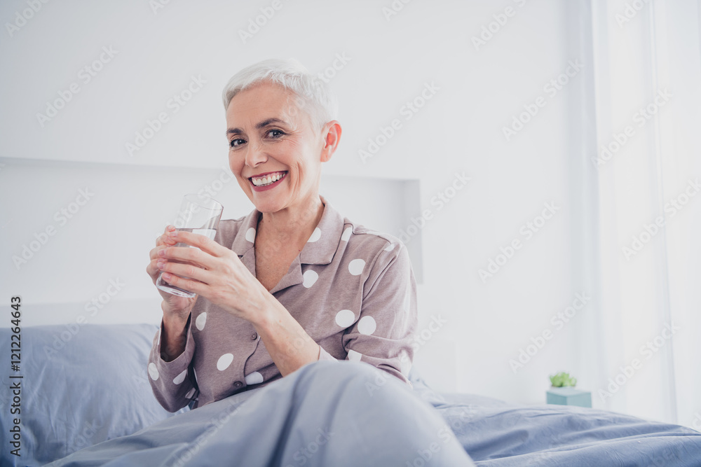 Charming grey-haired woman enjoying a refreshing morning in bed, holding a glass of water, exemplifying comfort and happiness Charming grey-haired woman enjoying a refreshing morning in bed, holding a glass of water, exemplifying comfort and happiness