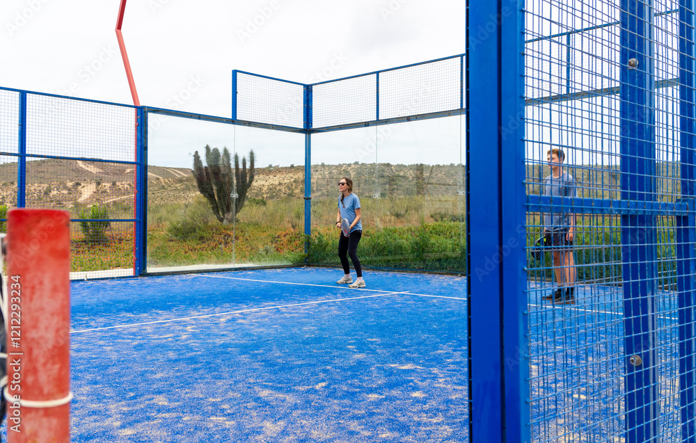 A view of a doubles match between a mother and her teenage son on a blue outdoor padel court, with a cloudy sky creating a moody atmosphere.
