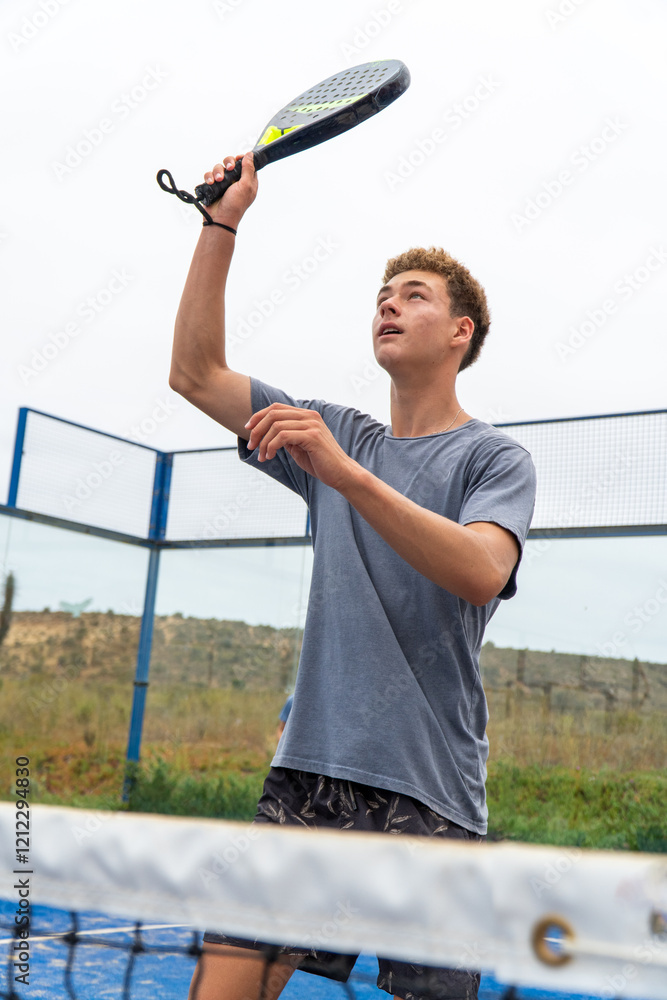 A teenager mid-action with a padel racket raised, poised to hit the ball during an intense game on the court.