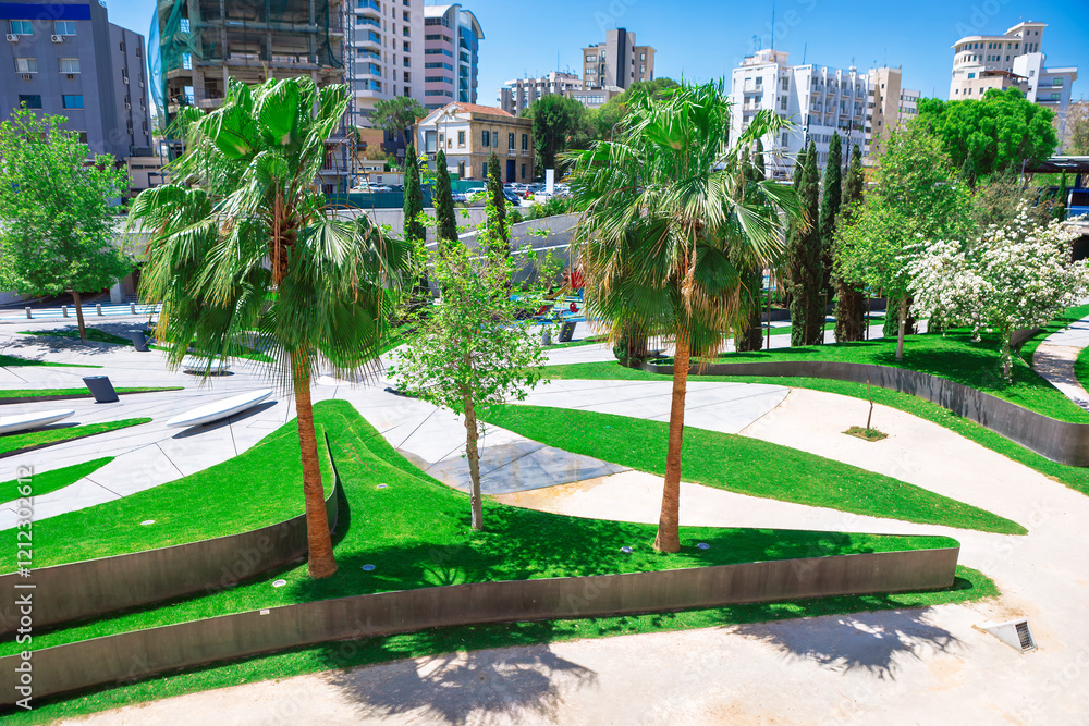 Modern urban park in Nicosia city, with green lawns, palm trees, and other vegetation, set against a backdrop of various buildings. Park features curving pathways and concrete structures